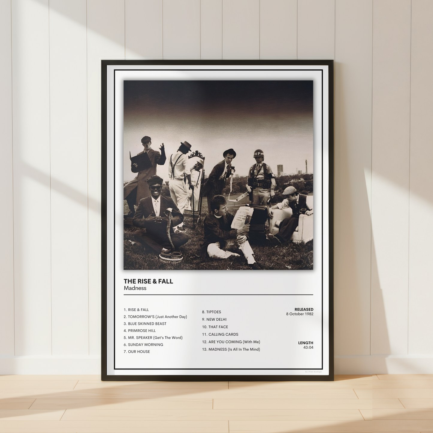 A framed black-and-white music poster for the band Madness 'The Rise & Fall' leaning against a white paneled wall on a light wood floor, with soft window shadows cast across the scene.