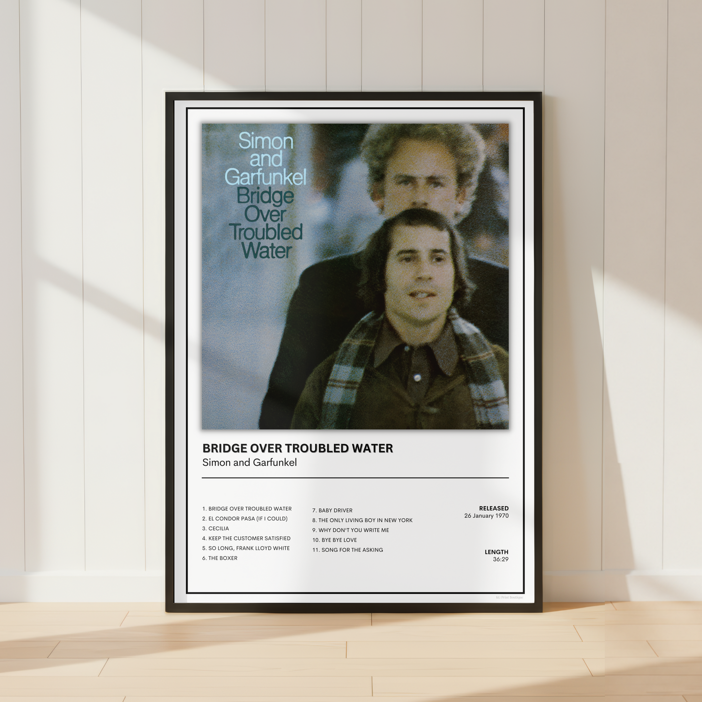 A framed music poster for Simon and Garfunkel's album 'Bridge Over Troubled Water' leaning against a white paneled wall on a light wood floor, with soft sunlight casting shadows.