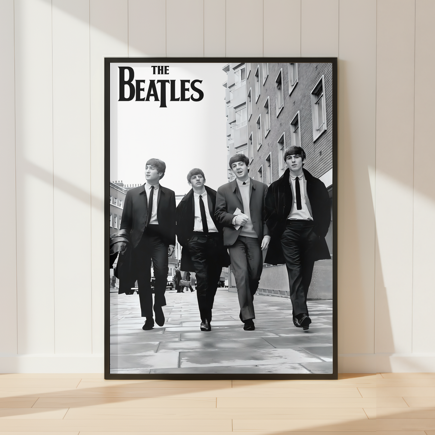 A framed black-and-white Beatles poster leaning against a white paneled wall on a light wood floor, with soft window shadows across the scene.