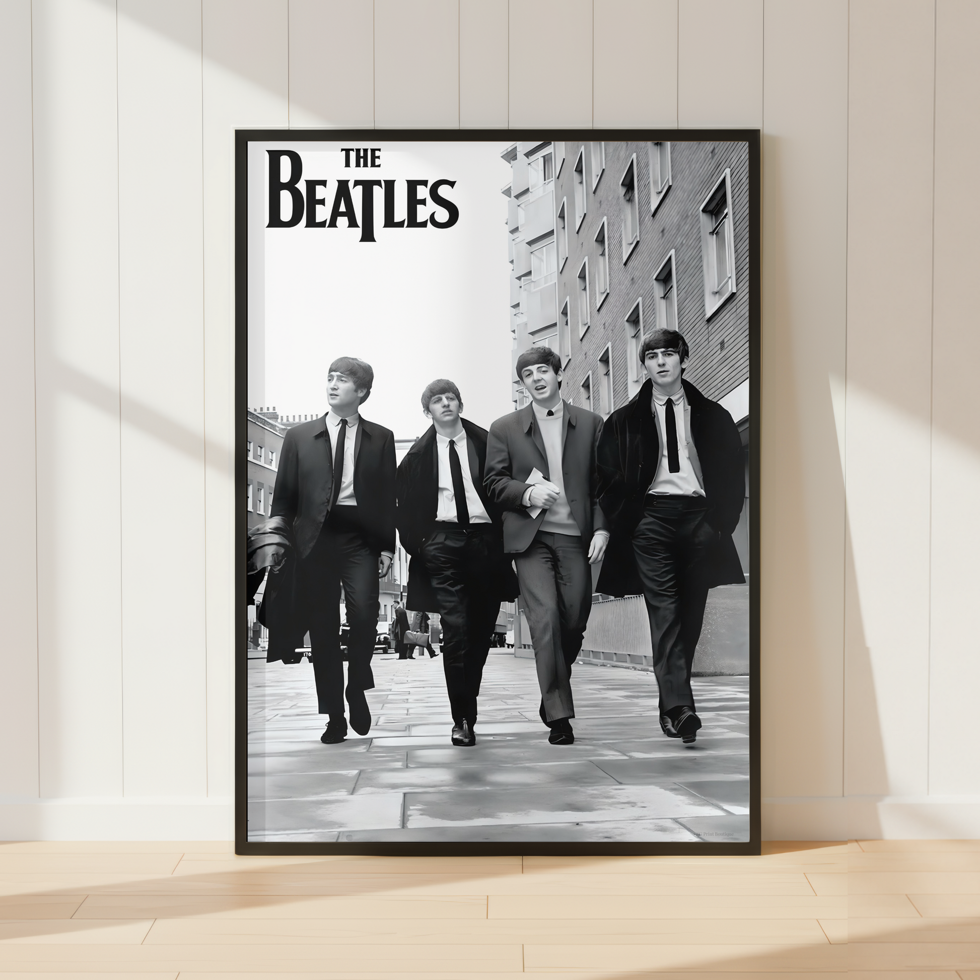 A framed black-and-white Beatles poster leaning against a white paneled wall on a light wood floor, with soft window shadows across the scene.
