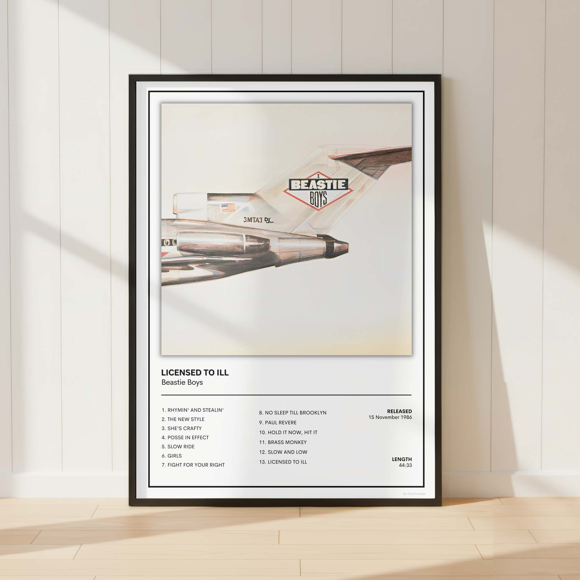 A framed music poster of U2's 'Beastie' leaning against a white paneled wall on a light wood floor, with soft sunlight casting shadows.