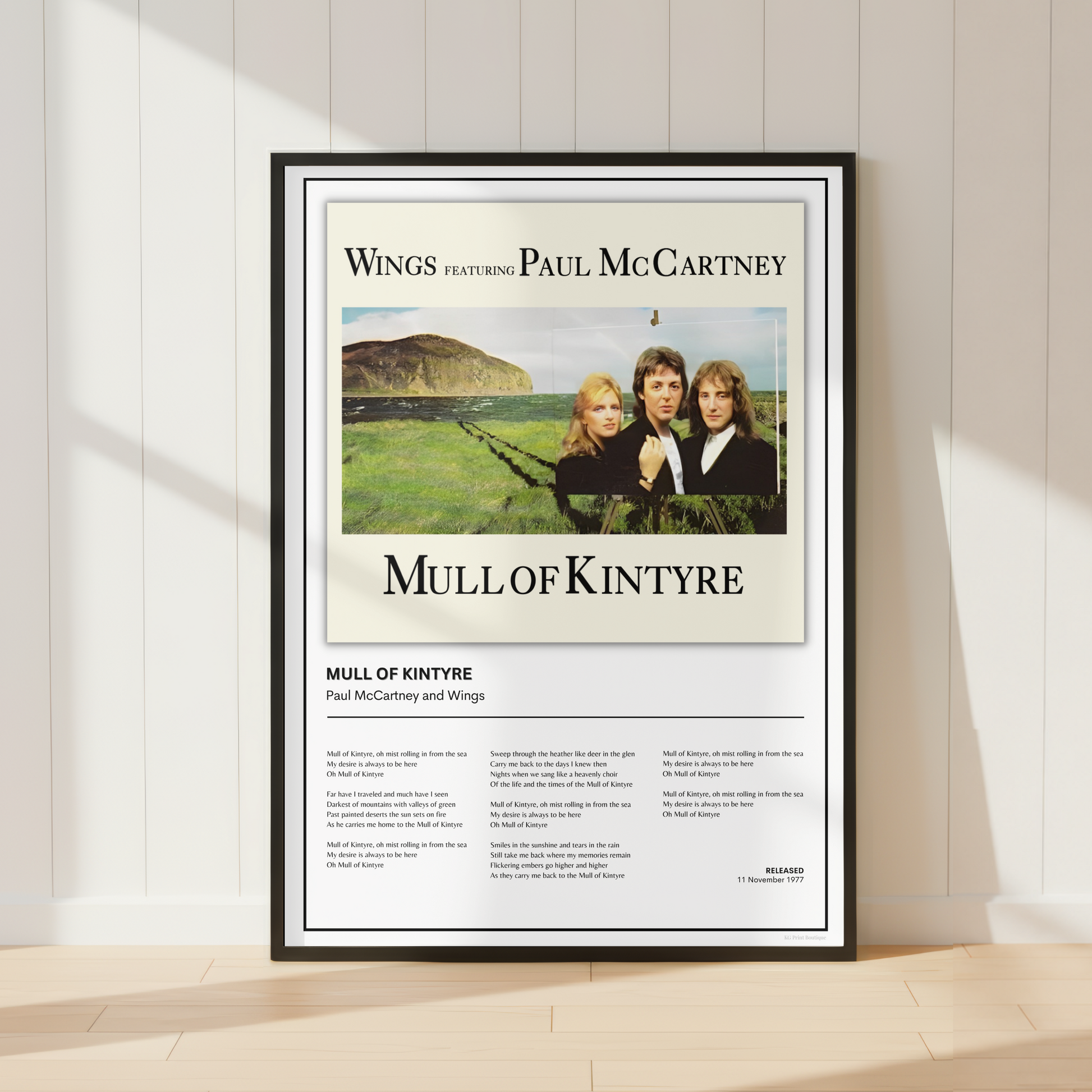 A framed Paul McCartney 'Mull of Kintyre' album poster print is displayed leaning against a white paneled wall on a light wood floor, with soft window shadows cast across the scene.
