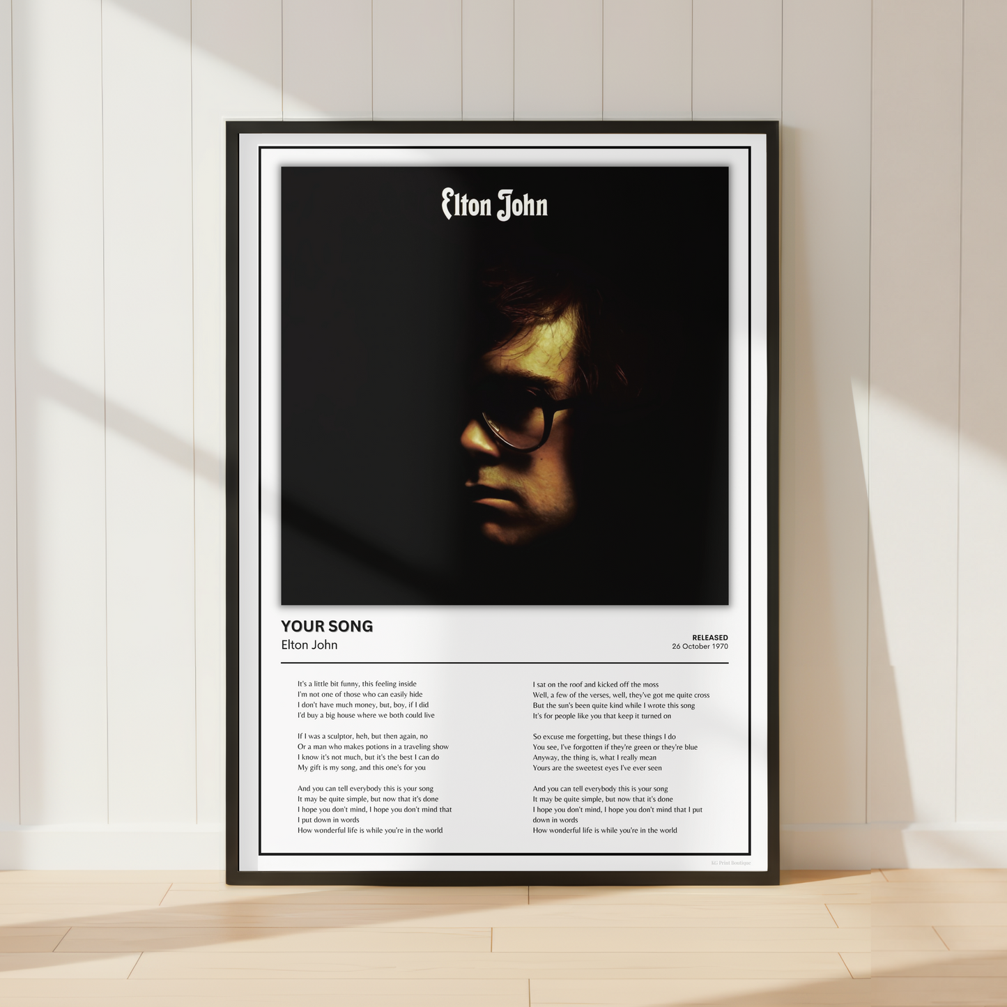 A framed Elton John song poster leaning against a white paneled wall on a light wood floor, with soft sunlight casting shadows across the scene.