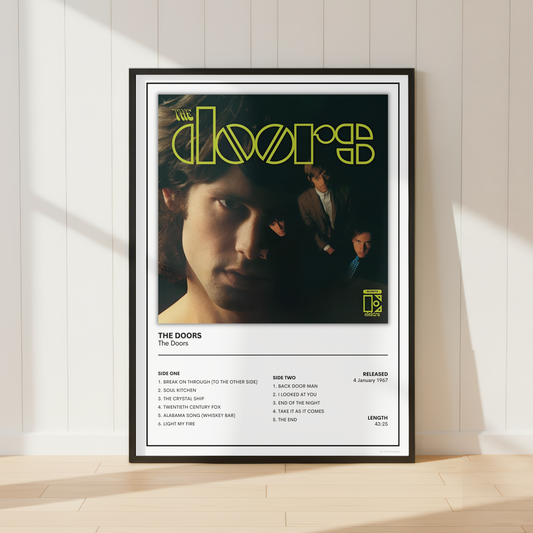 A framed The Doors 'The Doors' album poster is displayed leaning against a white paneled wall on a light wood floor, with soft sunlight casting shadows.