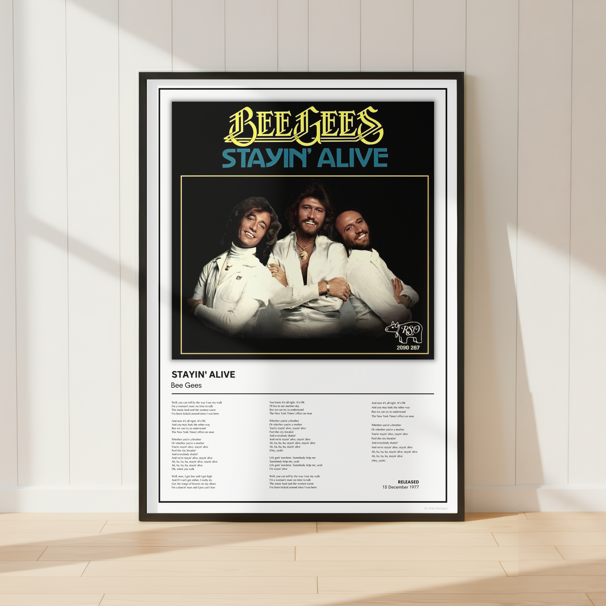 A framed Bee Gees 'Stayin' Alive' poster leaning against a white paneled wall on a light wood floor, with soft sunlight casting shadows across the scene.