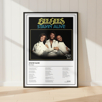 A framed Bee Gees 'Stayin' Alive' poster leaning against a white paneled wall on a light wood floor, with soft sunlight casting shadows across the scene.
