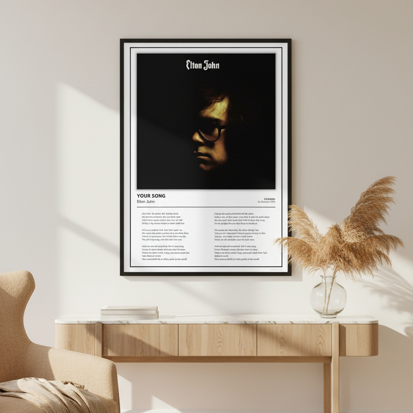 A framed Elton John music poster displayed on a light wood wall and floor. The poster features a black-and-white portrait of Elton John with the text 'Elton John' at the top and 'YOUR SONG' with song titles and lyrics below.