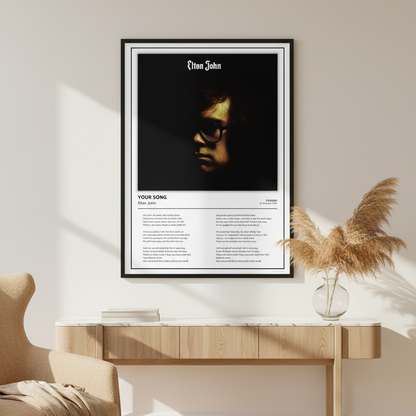 A framed Elton John music poster displayed on a light wood wall and floor. The poster features a black-and-white portrait of Elton John with the text 'Elton John' at the top and 'YOUR SONG' with song titles and lyrics below.