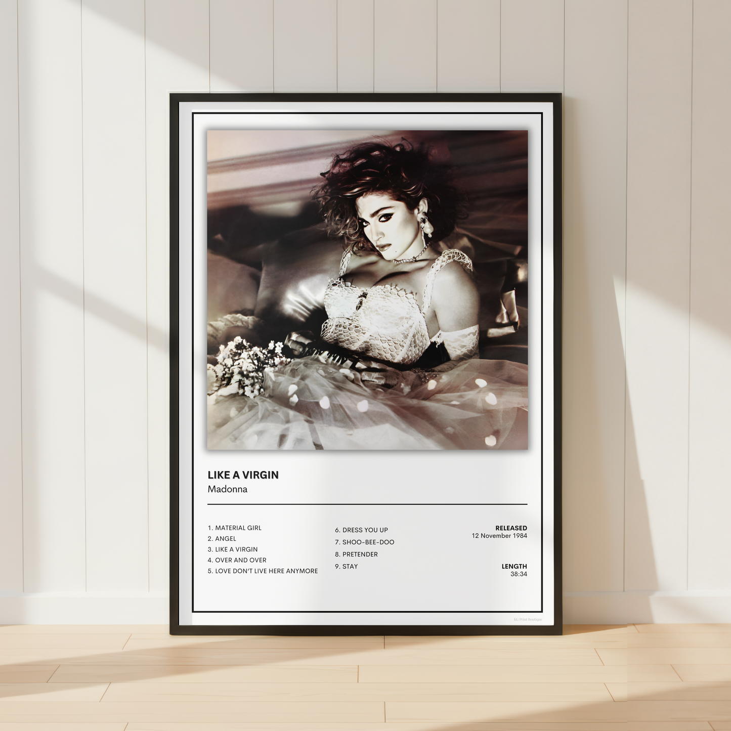 A framed music poster of Madonna's 'Like a Virgin' leaning against a white paneled wall on a light wood floor, with soft sunlight casting shadows.