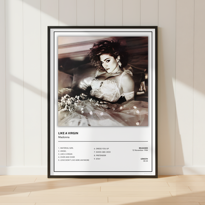 A framed music poster of Madonna's 'Like a Virgin' leaning against a white paneled wall on a light wood floor, with soft sunlight casting shadows.