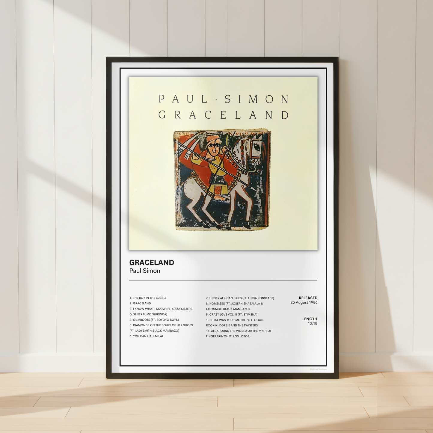 A framed music poster of Paul Simon & Graceland leaning against a white paneled wall on a light wood floor, with soft sunlight casting shadows.