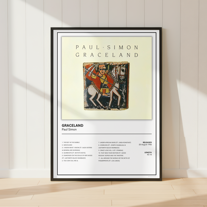 A framed music poster of Paul Simon & Graceland leaning against a white paneled wall on a light wood floor, with soft sunlight casting shadows.