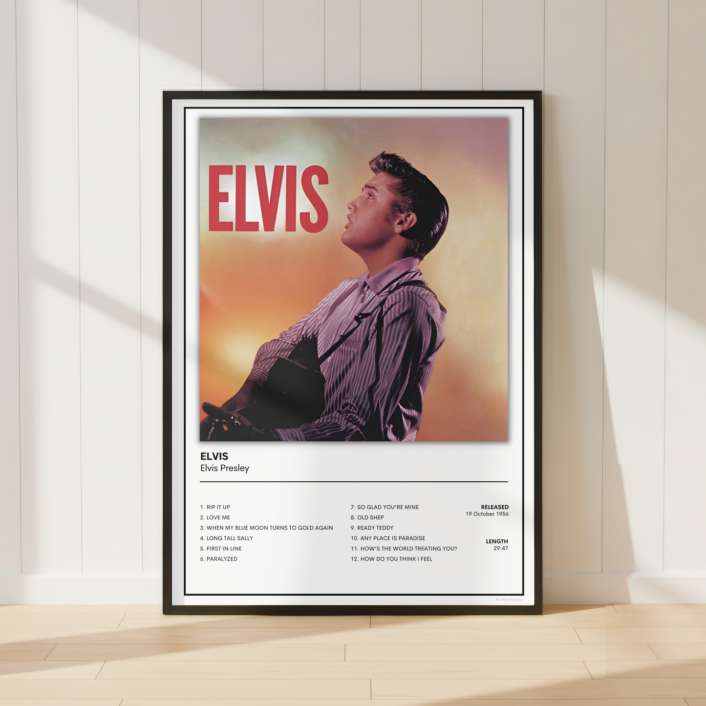A framed Elvis Presley poster leaning against a white paneled wall on a light wood floor, with warm sunlight casting shadows across the scene.