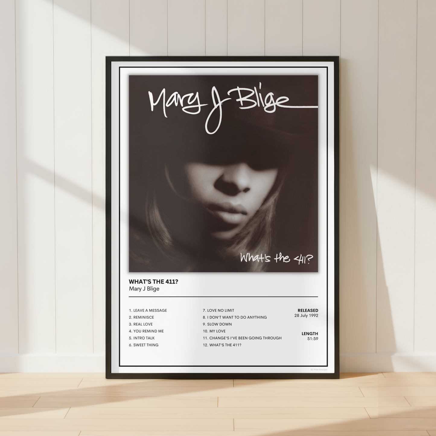 A framed black and white music poster of Mary J. Blige's album 'What's The 417?' leaning against a white paneled wall on a light wood floor.