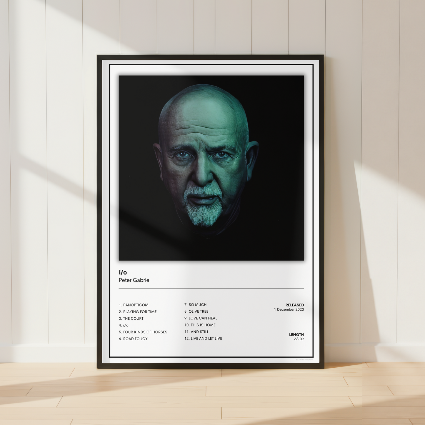 A framed music poster of Peter Gabriel's album 'Life' leaning against a white paneled wall on a light wood floor, with soft window shadows.