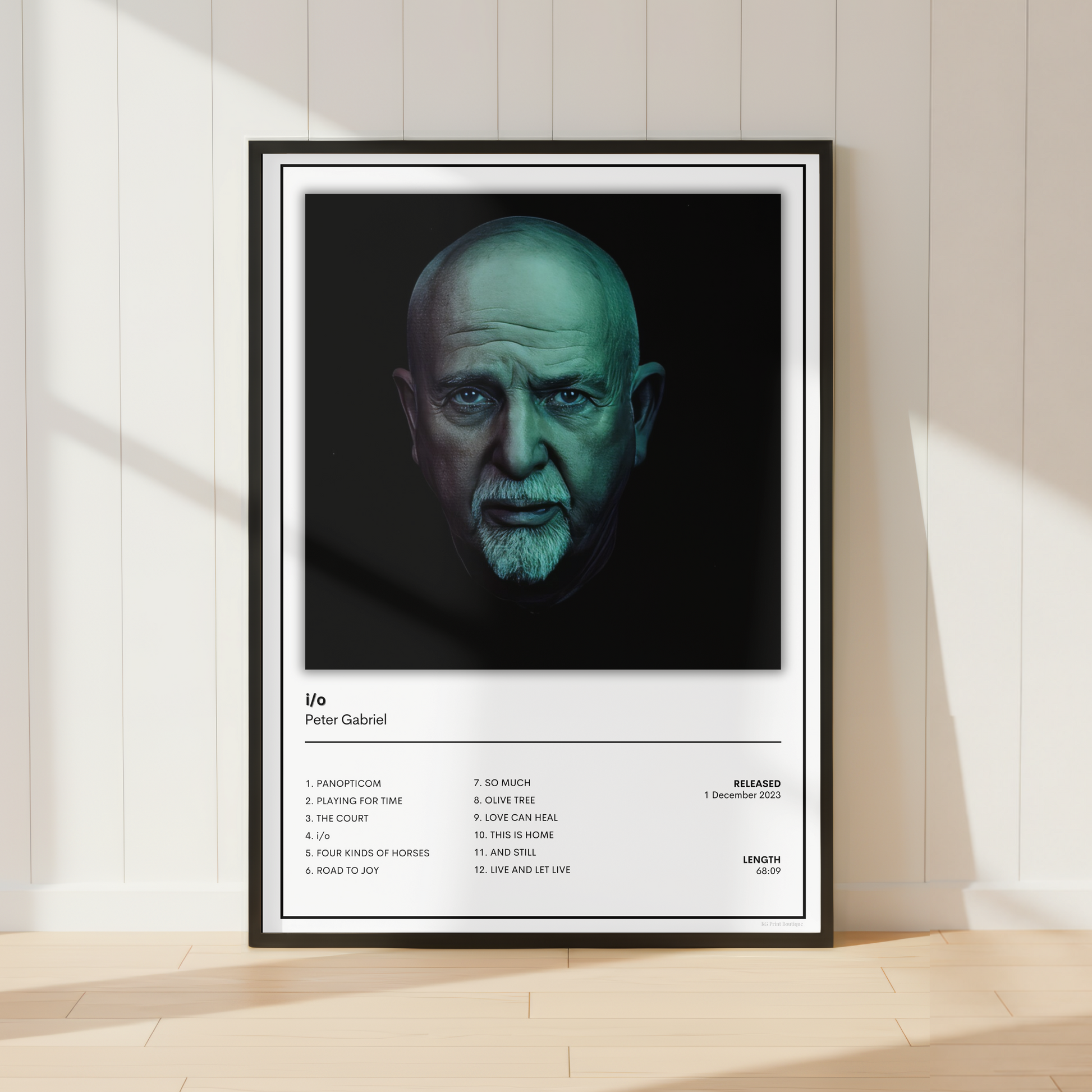 A framed music poster of Peter Gabriel's album 'Life' leaning against a white paneled wall on a light wood floor, with soft window shadows.