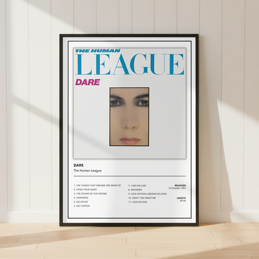 A framed music poster for 'Dare' by The Human League, styled leaning against a white paneled wall on a light wood floor with soft window shadows.
