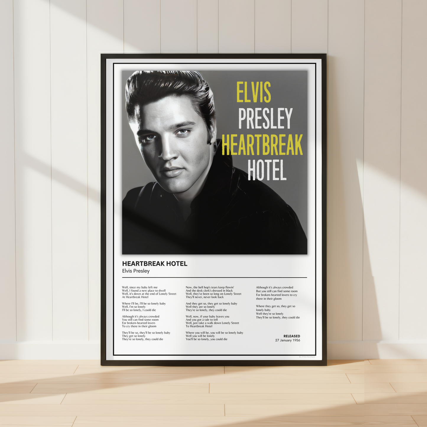 A framed vertical poster of Elvis Presley's 'Heartbreak Hotel' leaning against a white paneled wall on a light wood floor, with soft window shadows cast across the scene.
