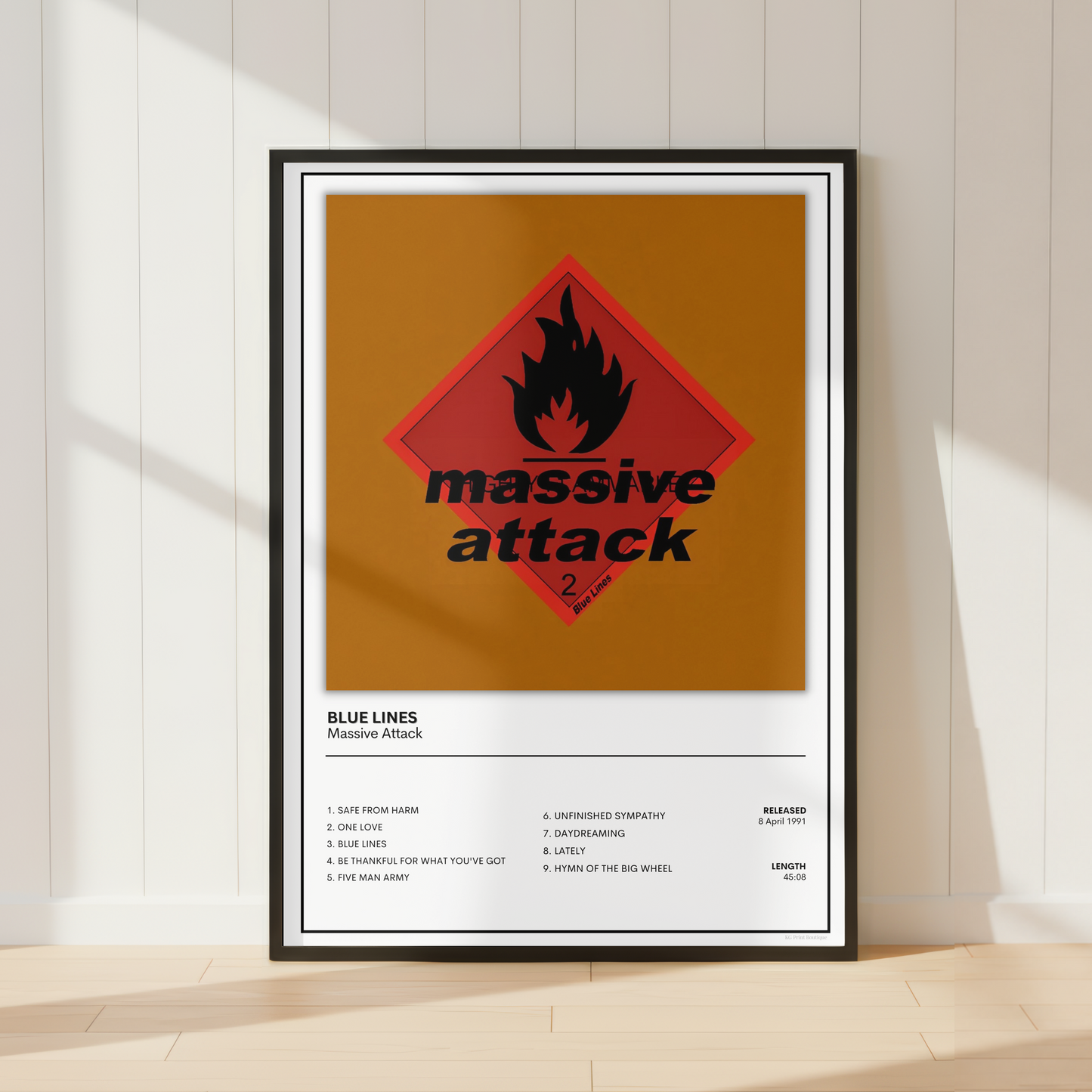 A framed poster leaning against a white paneled wall on a light wood floor. The poster features a vintage-style hazard sign with a red diamond, black flame, and the words 'massive attack' on an orange-brown background, with additional text below reading 'BLUE LINES' and 'Massive Attack.