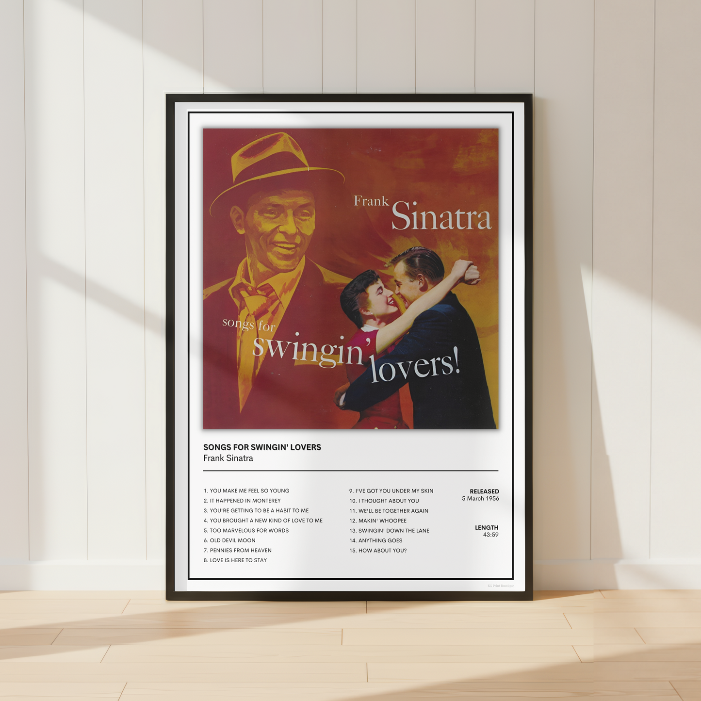 A framed Frank Sinatra 'Swingin' Lovers' music poster is displayed leaning against a white paneled wall on a light wood floor, with soft sunlight casting shadows.