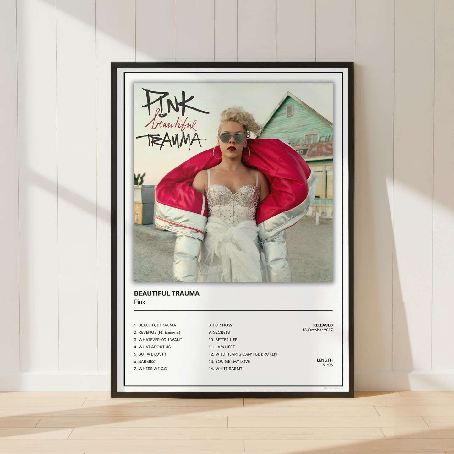 A framed music poster of Pink's album 'Beautiful Trauma' leaning against a white paneled wall on a light wood floor. The poster features a photo of Pink in a white corset and a bright pink jacket, with the album title and track list printed below.