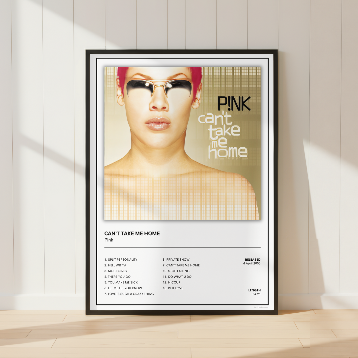 A framed music poster of Pink's 'Can't Take Me Home' leaning against a white paneled wall on a light wood floor, with soft window shadows cast across the scene.