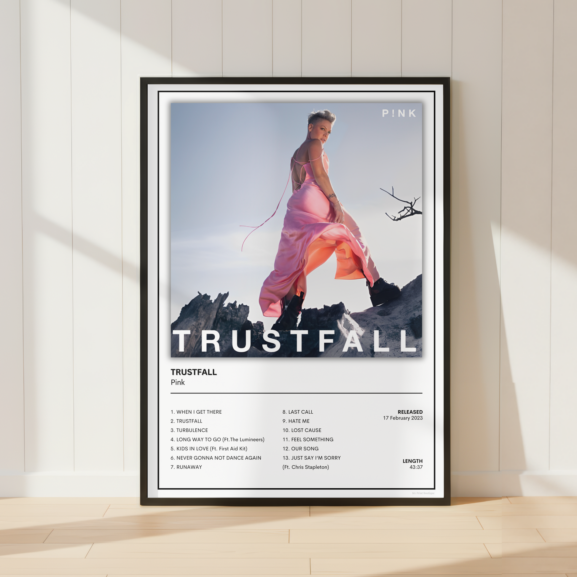 A framed music poster for Pink's album 'Trustfall' leaning against a white paneled wall on a light wood floor. The poster features a woman in a pink dress on a mountain with a helicopter, and the album title and track list printed below.
