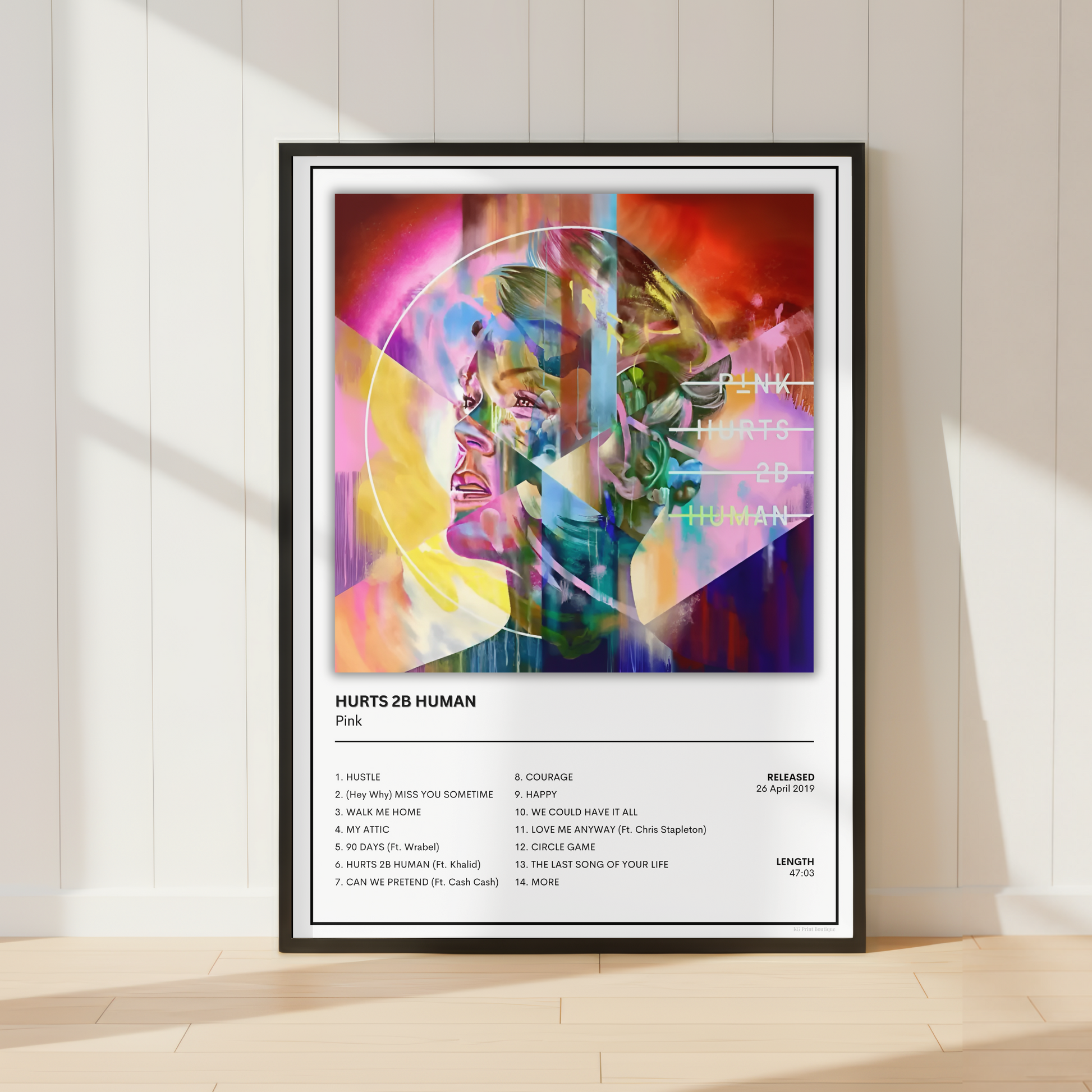 A framed music poster for Pink's album 'HURTS 28 HUMAN' leaning against a white paneled wall on a light wood floor, with colorful abstract artwork and tracklist details.