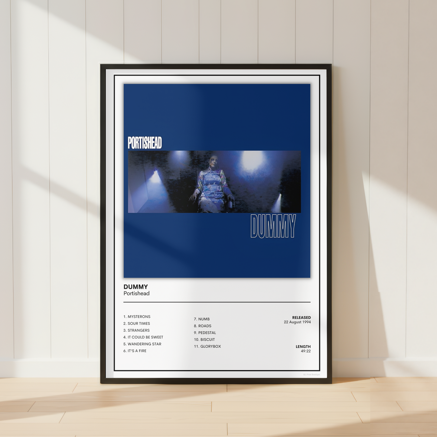 A framed music poster for the song 'Dummy' by Portishead, leaning against a white paneled wall on a light wood floor. The poster features a dark blue top section with the band name and a moody, low-key image of a person, and a white bottom section with track listing details.