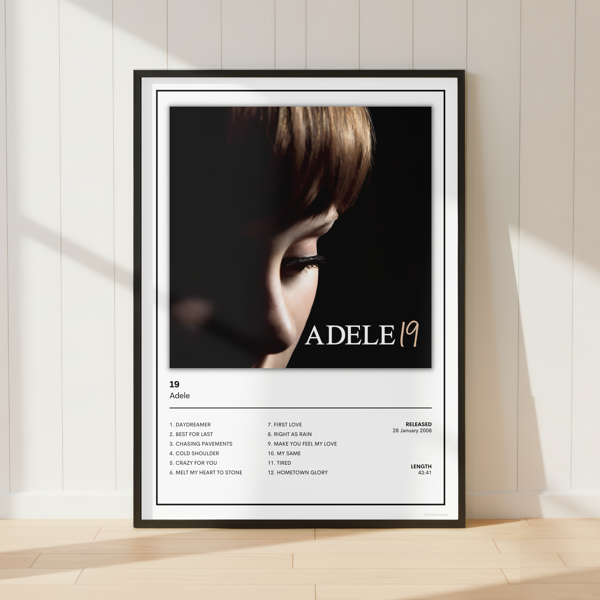 A framed Adele 19 album poster leaning against a white paneled wall on a light wood floor, with soft sunlight casting shadows across the scene.