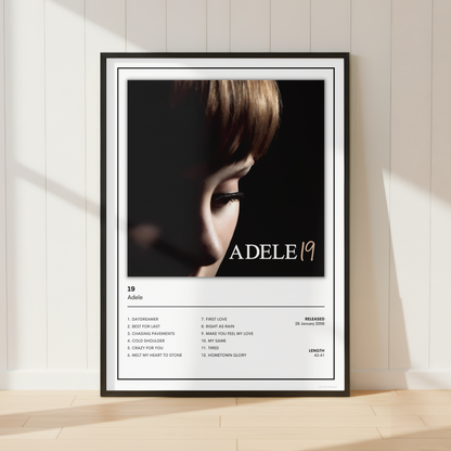 A framed Adele 19 album poster leaning against a white paneled wall on a light wood floor, with soft sunlight casting shadows across the scene.