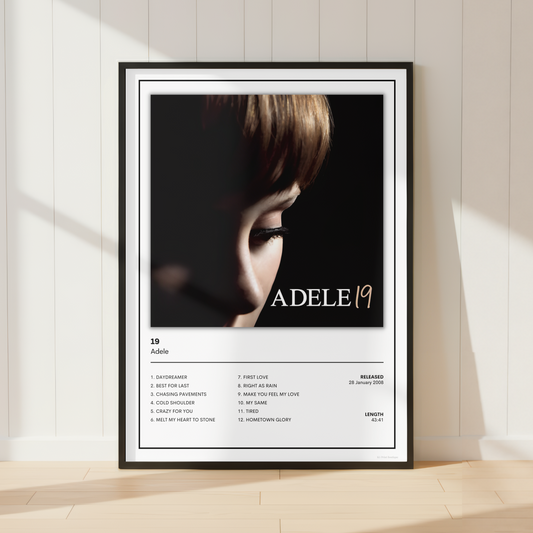 A framed Adele 19 album poster leaning against a white paneled wall on a light wood floor, with soft sunlight casting shadows across the scene.