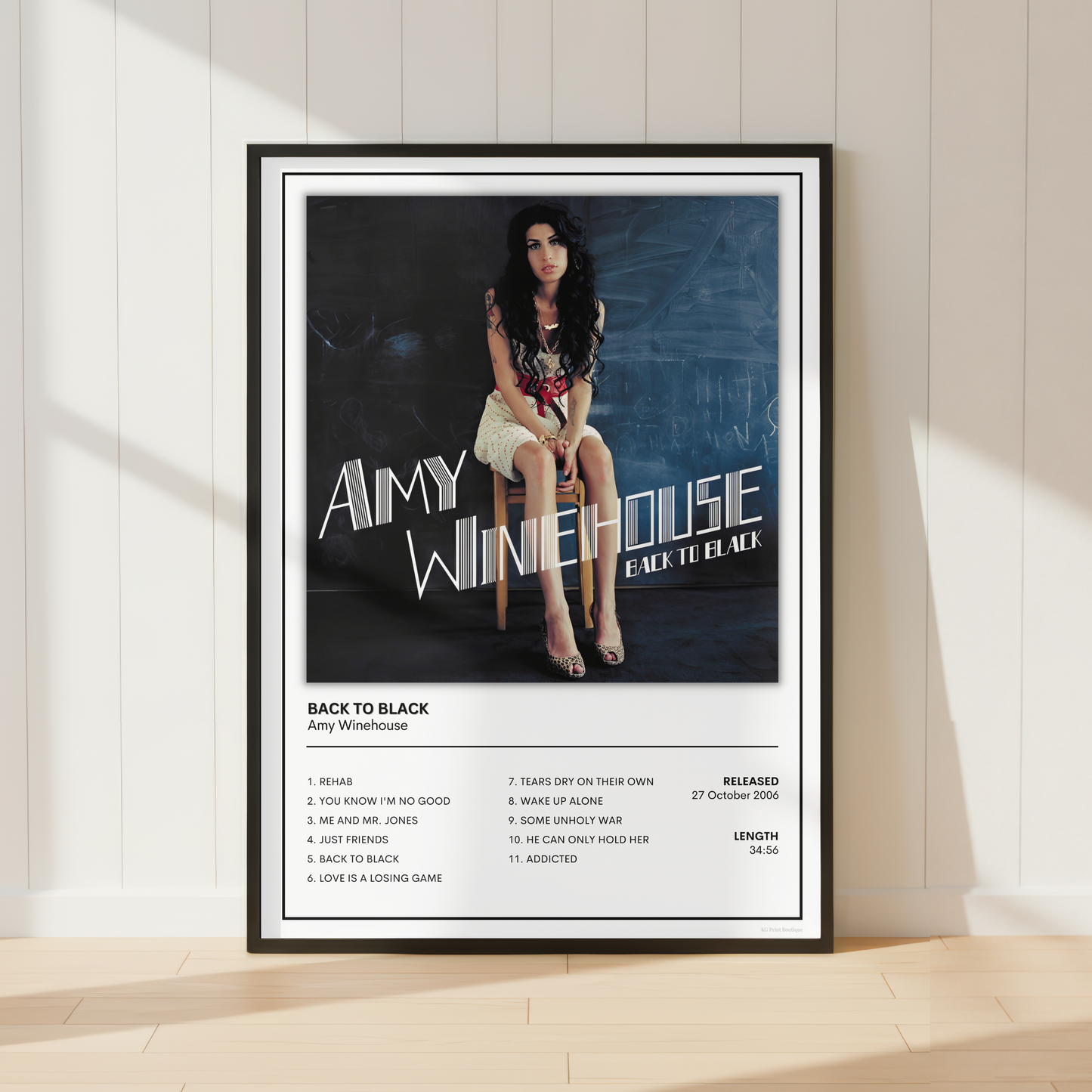 A framed music poster of Amy Winehouse's album 'Back to Black' leaning against a white paneled wall on a light wood floor.