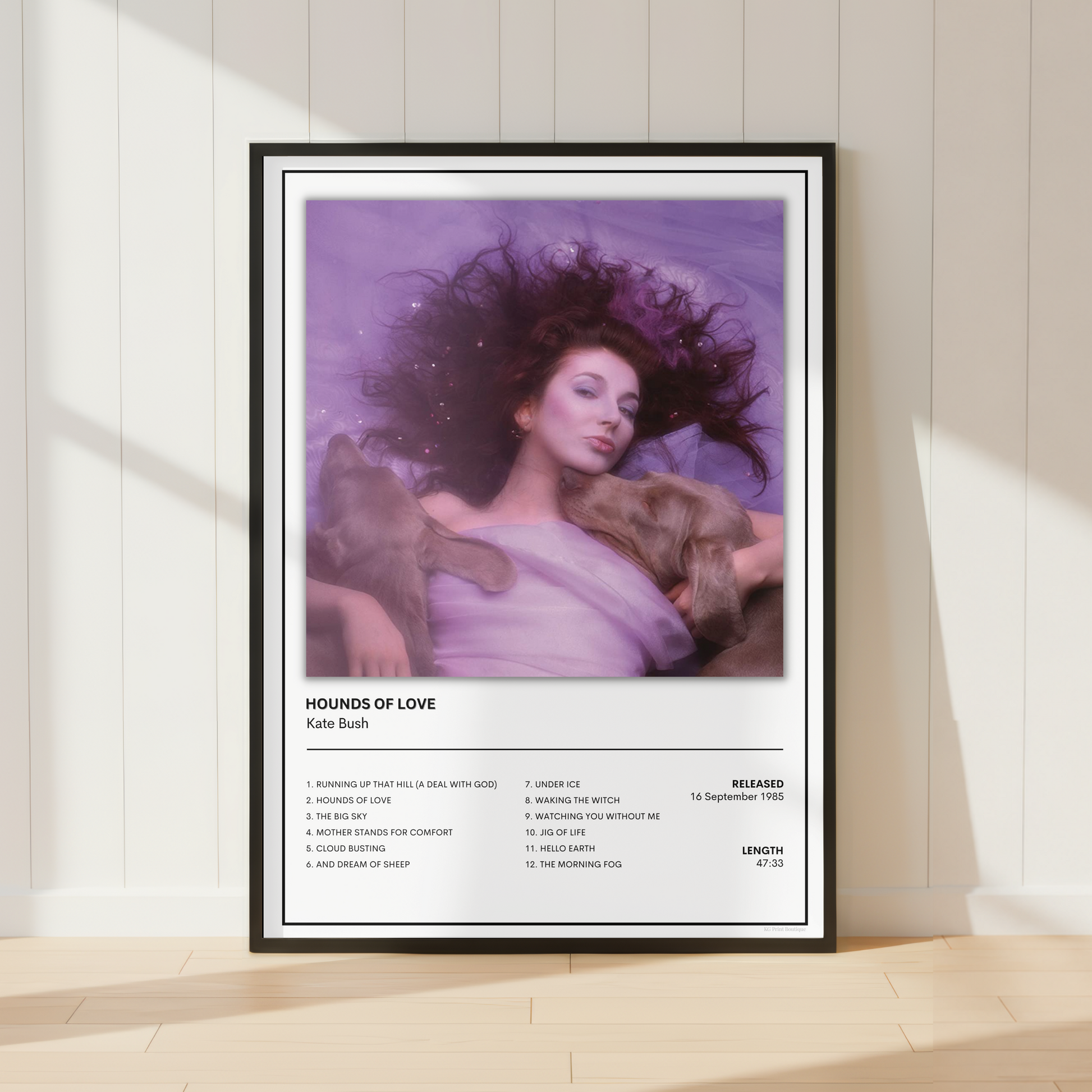 A framed music poster of Kate Bush's 'Hounds of Love' leaning against a white paneled wall on a light wood floor, styled with soft window shadows.