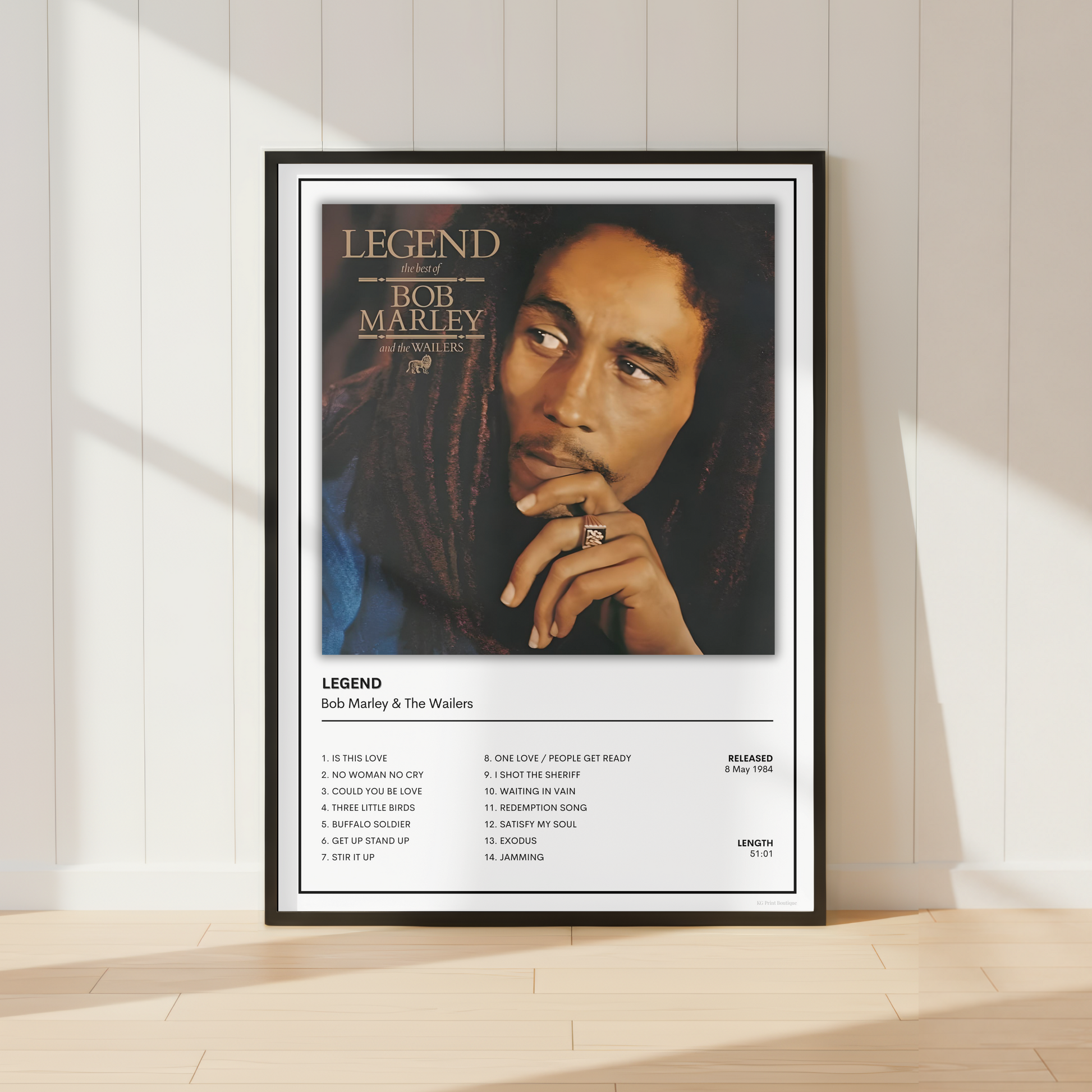 A framed Bob Marley 'Legend' music poster is displayed on a light wood floor against a white paneled wall with soft window shadows.
