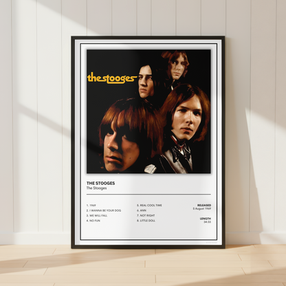 A framed music poster of the band The Stooges leaning against a white paneled wall on a light wood floor. The poster features a black background with the band members' portrait, the yellow 'the stooges' logo, and track listing text below.