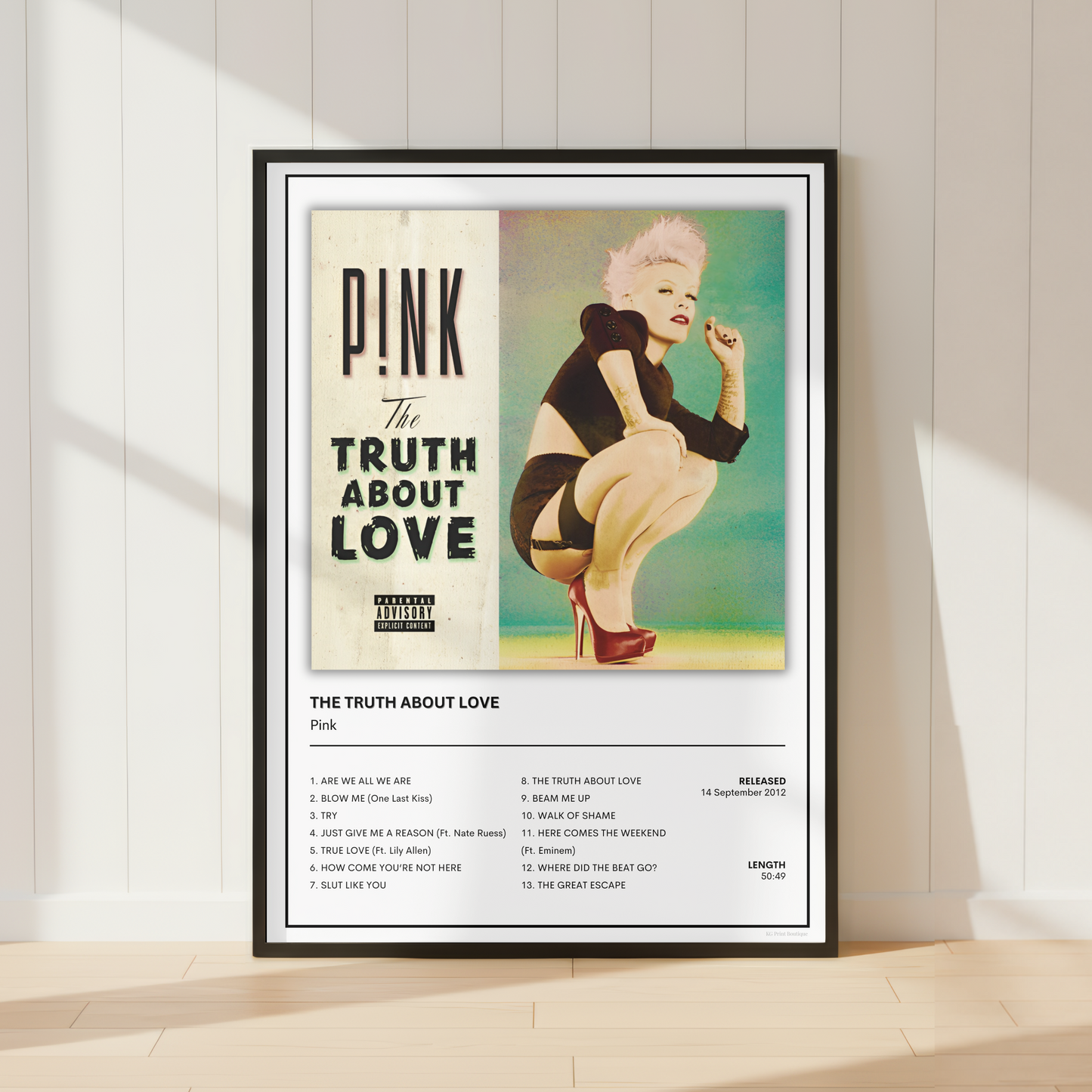 A framed Pink 'The Truth About Love' music poster leaning against a white paneled wall on a light wood floor, with soft diagonal shadows across the scene.