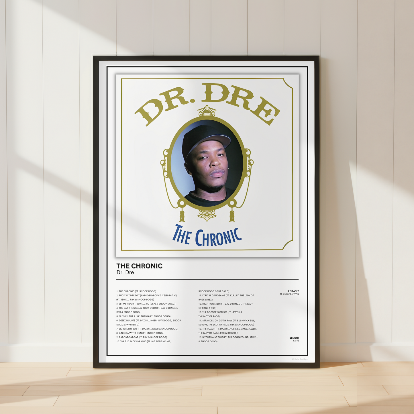A framed music poster of Dr. Dre's 'The Chronic' leaning against a white paneled wall on a light wood floor, with soft diagonal sunlight casting shadows.