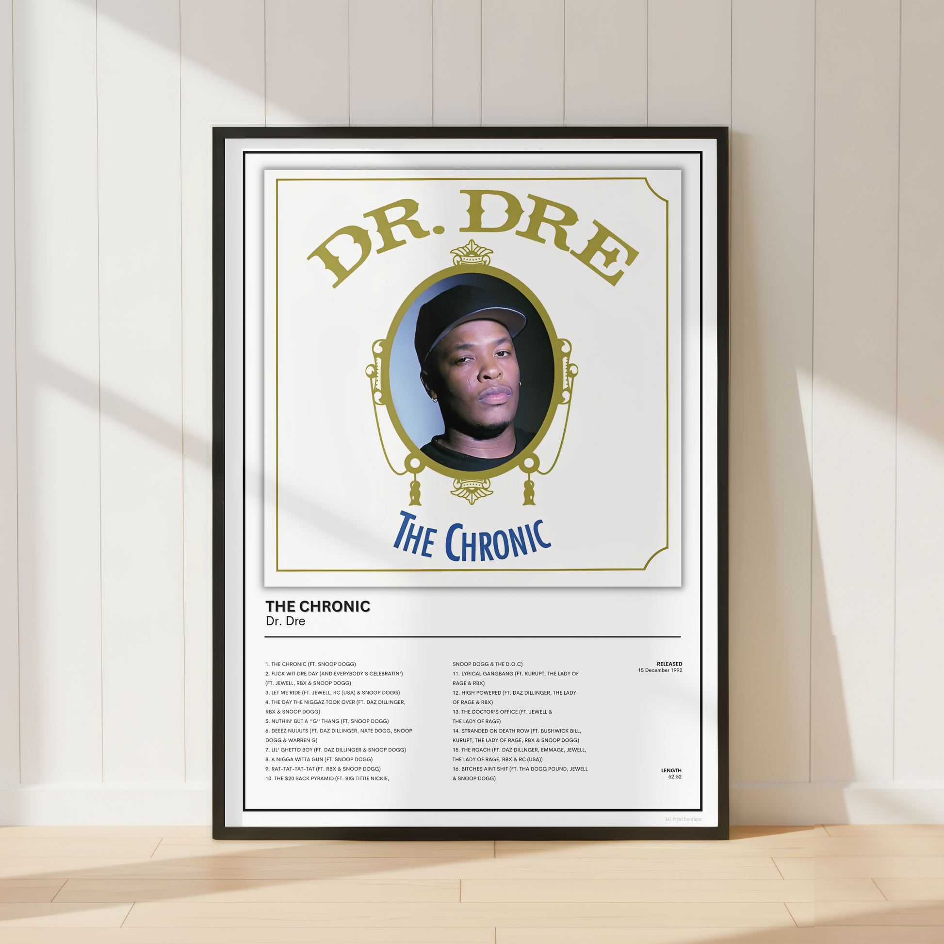 A framed music poster of Dr. Dre's 'The Chronic' leaning against a white paneled wall on a light wood floor, with soft diagonal sunlight casting shadows.