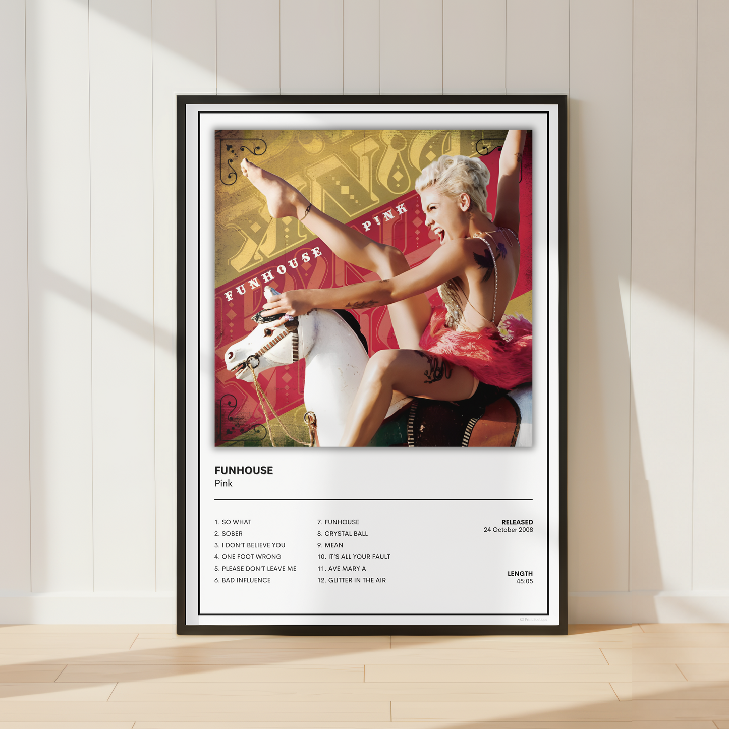 A framed music poster for Pink's album 'Funhouse' leaning against a white paneled wall on a light wood floor. The poster features a colorful album-cover photo of Pink on a carousel horse above a printed tracklist and release details.