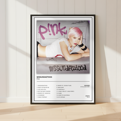 A framed music poster of Pink's 'Missundaztood' leaning against a white paneled wall on a light wood floor, with soft sunlight casting shadows.