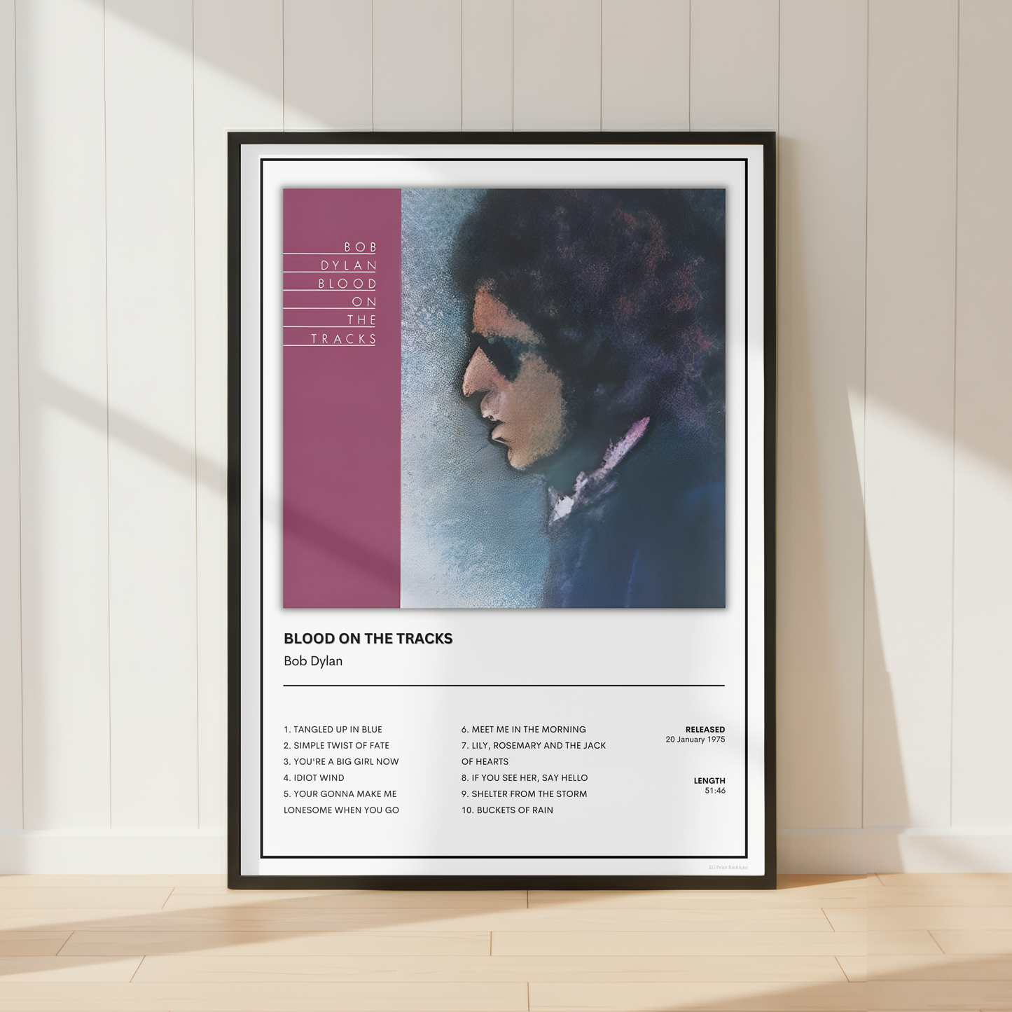 A framed music poster for Bob Dylan's album 'Blood on the Tracks' leaning against a white paneled wall on a light wood floor, with soft window shadows.