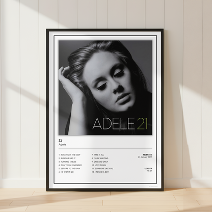 A framed black-and-white music poster of Adele's album '21' leaning against a white paneled wall on a light wood floor, with soft window shadows cast across the scene.