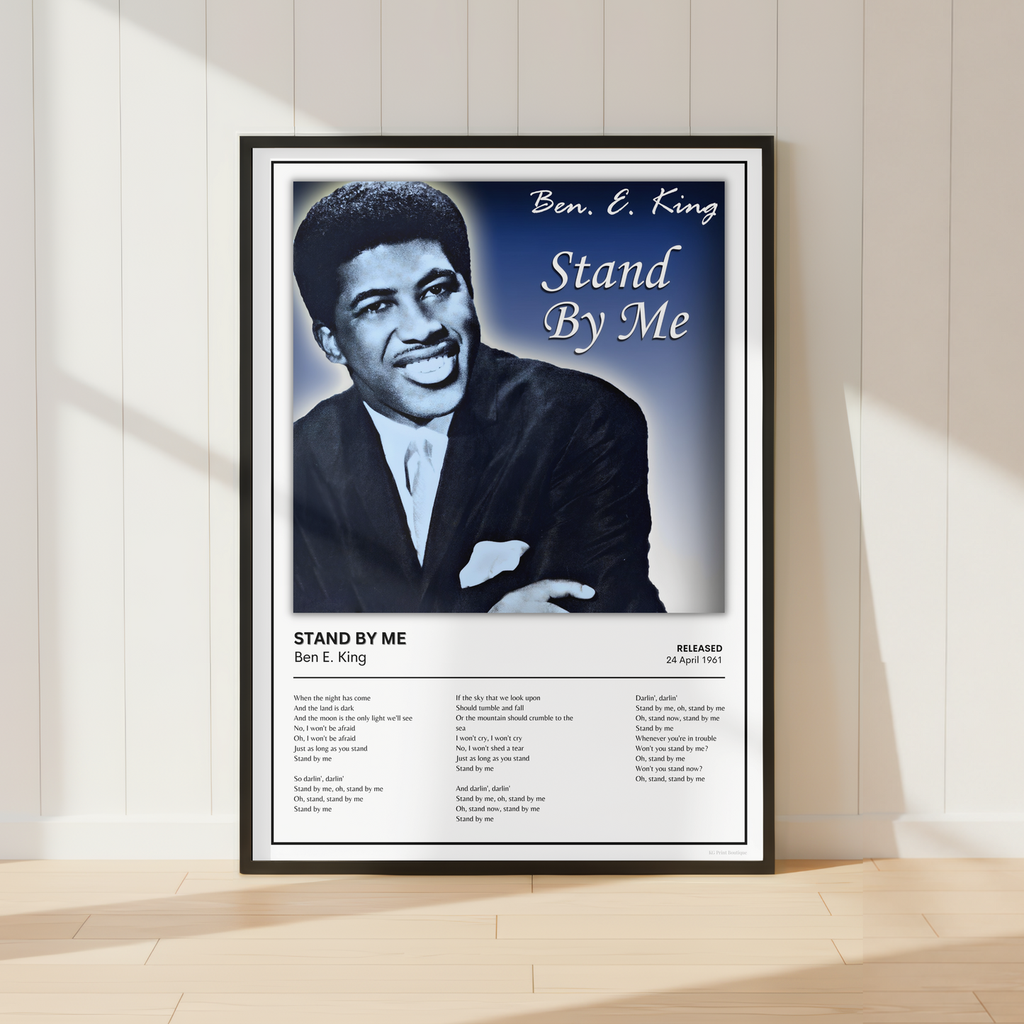 A framed music poster of Ben E. King's 'Stand By Me' leaning against a white paneled wall on a light wood floor, with soft sunlight casting shadows.