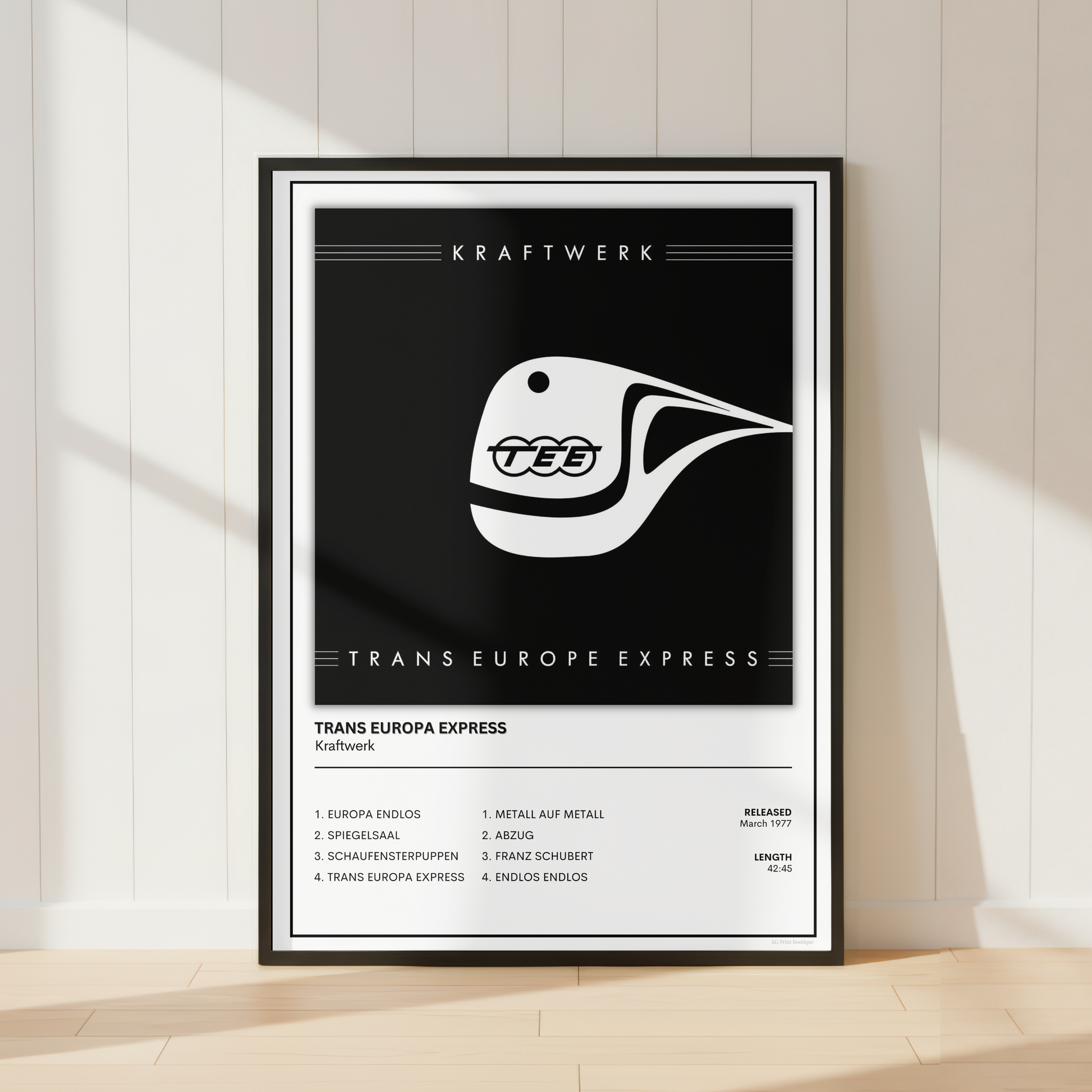 A framed black and white poster featuring the Trans Europe Express train logo and route text, leaning against a white paneled wall on a light wood floor.