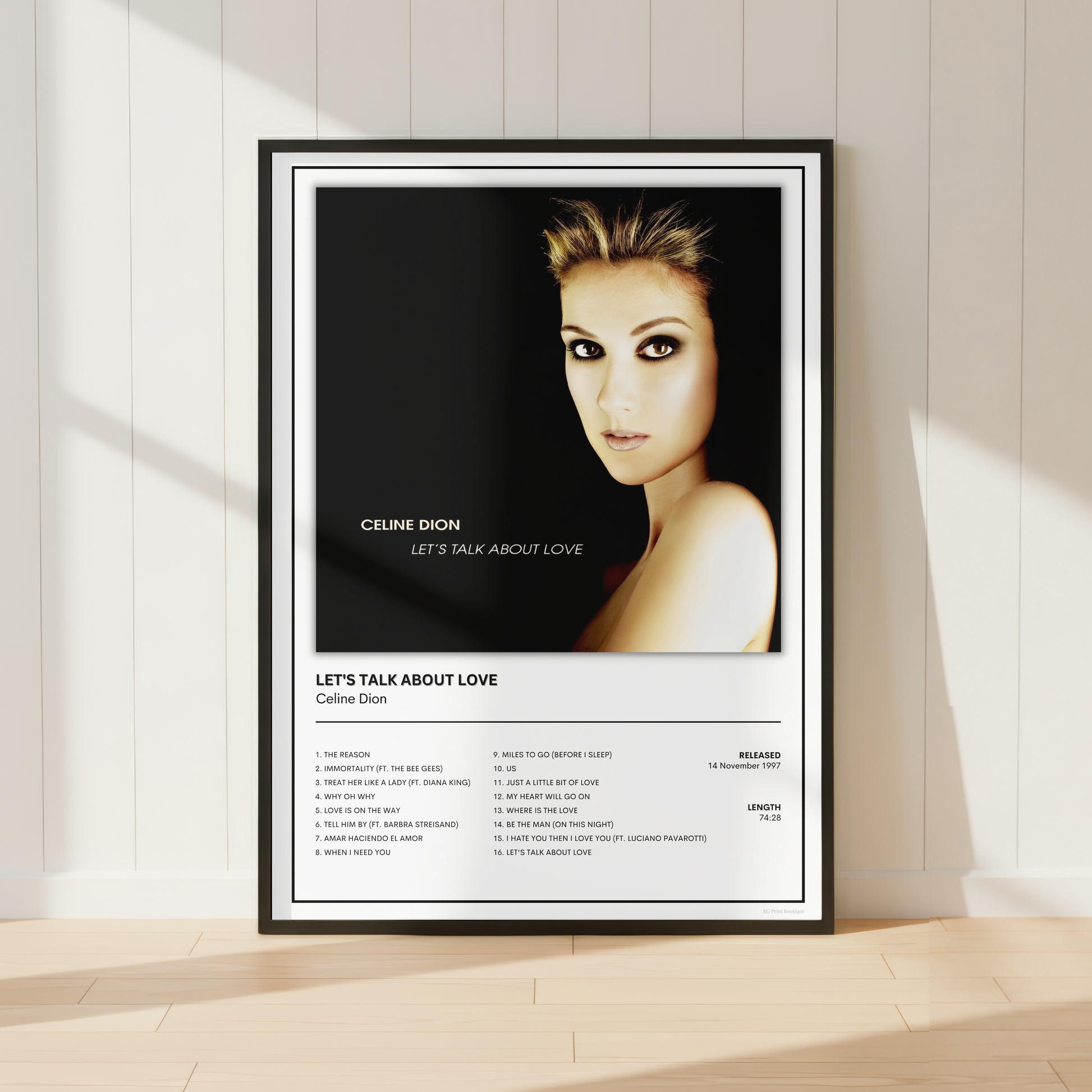 A framed music poster of Celine Dion's 'Let's Talk About Love' leaning against a white paneled wall on a light wood floor.