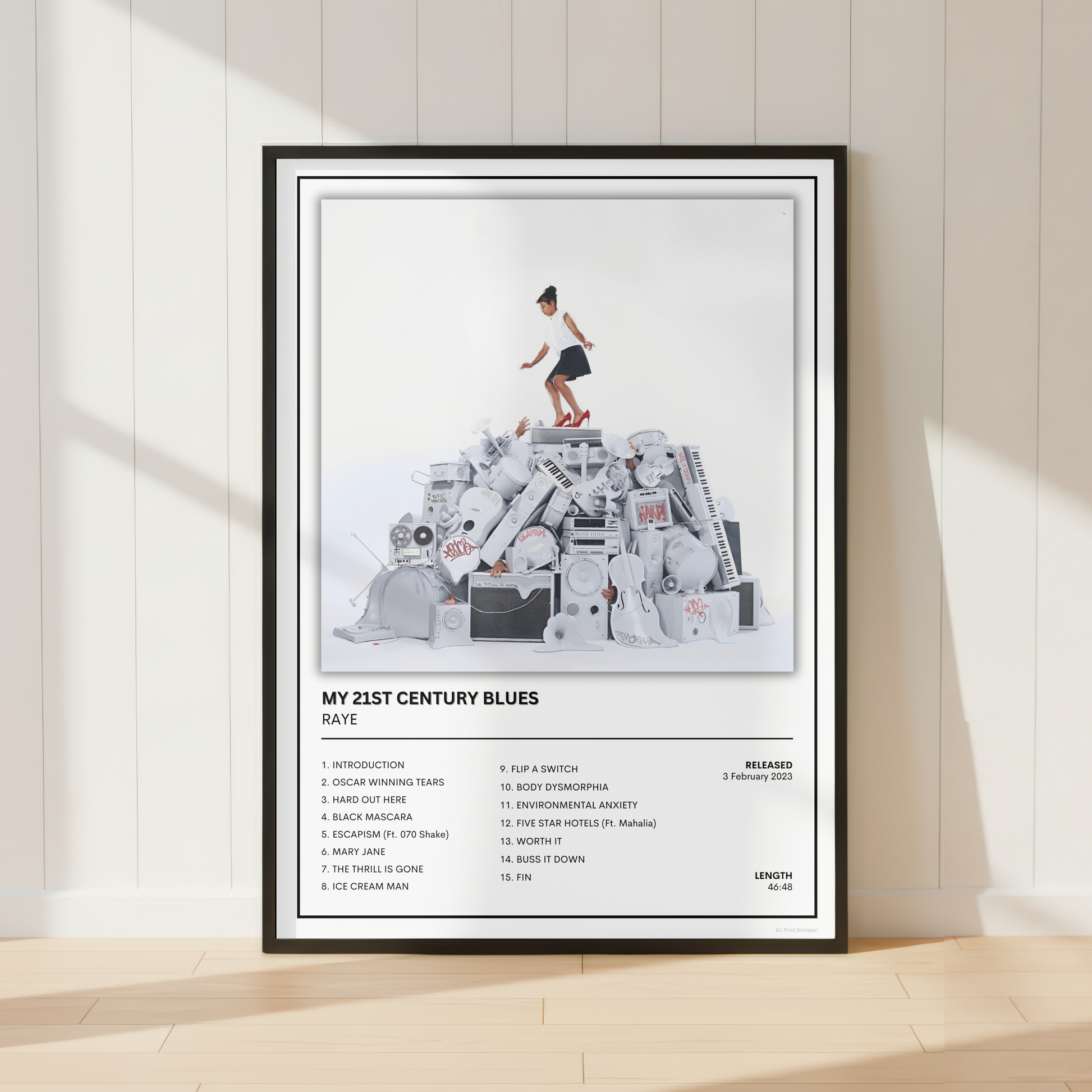 A framed music poster for the band RAYE's album 'My 21st Century Blues' is displayed leaning against a white paneled wall on a light wood floor. The poster features the album cover artwork with a child standing on a pile of cassette tapes, tracklist text, and the Muse logo.