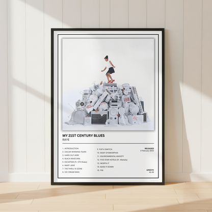 A framed music poster for the band RAYE's album 'My 21st Century Blues' is displayed leaning against a white paneled wall on a light wood floor. The poster features the album cover artwork with a child standing on a pile of cassette tapes, tracklist text, and the Muse logo.