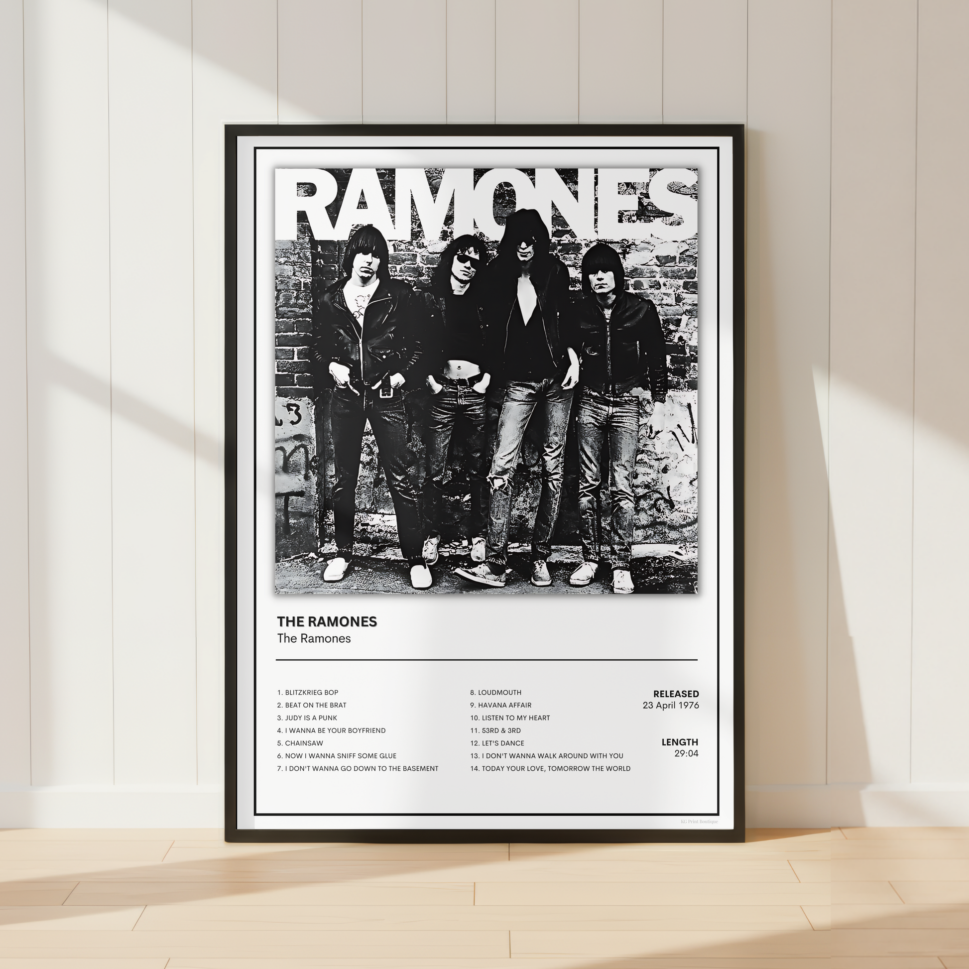 A framed black and white Ramones poster leaning against a white paneled wall on a light wood floor, with soft sunlight casting shadows across the scene.
