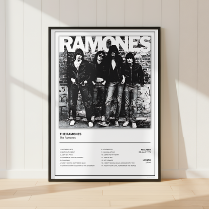 A framed black and white Ramones poster leaning against a white paneled wall on a light wood floor, with soft sunlight casting shadows across the scene.